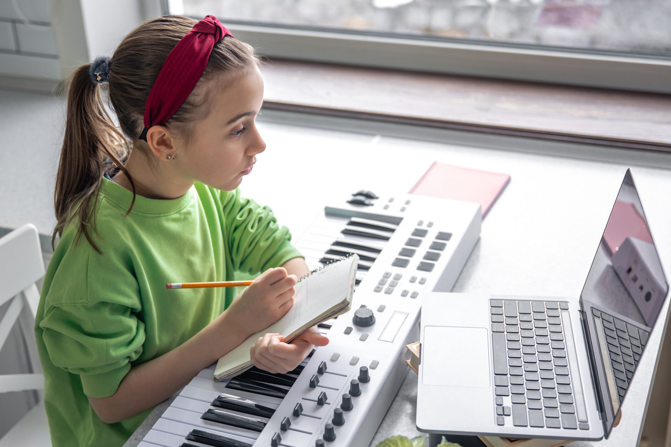 Ein junges Mädchen sitzt mit einem Notizblock an einem MIDI-Keyboard vor einem Laptop und komponiert Musik im digitalen Unterricht mit groove2grow.