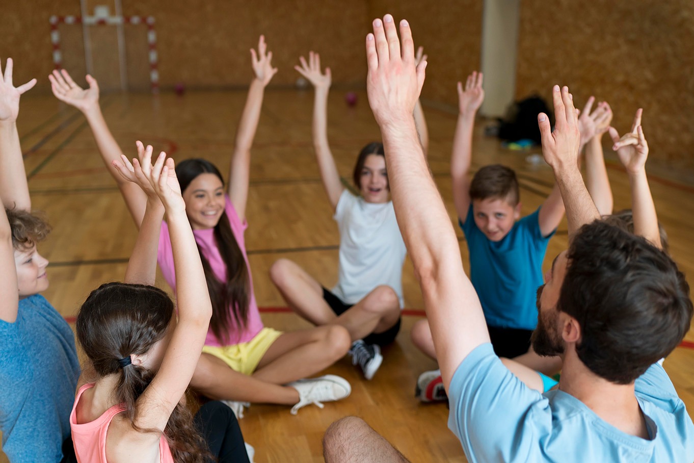 Eine Gruppe von Kindern und ein Lehrer im Sitzkreis strecken konzentriert ihre Hände nach oben – Training der Feinmotorik durch Tanz.
