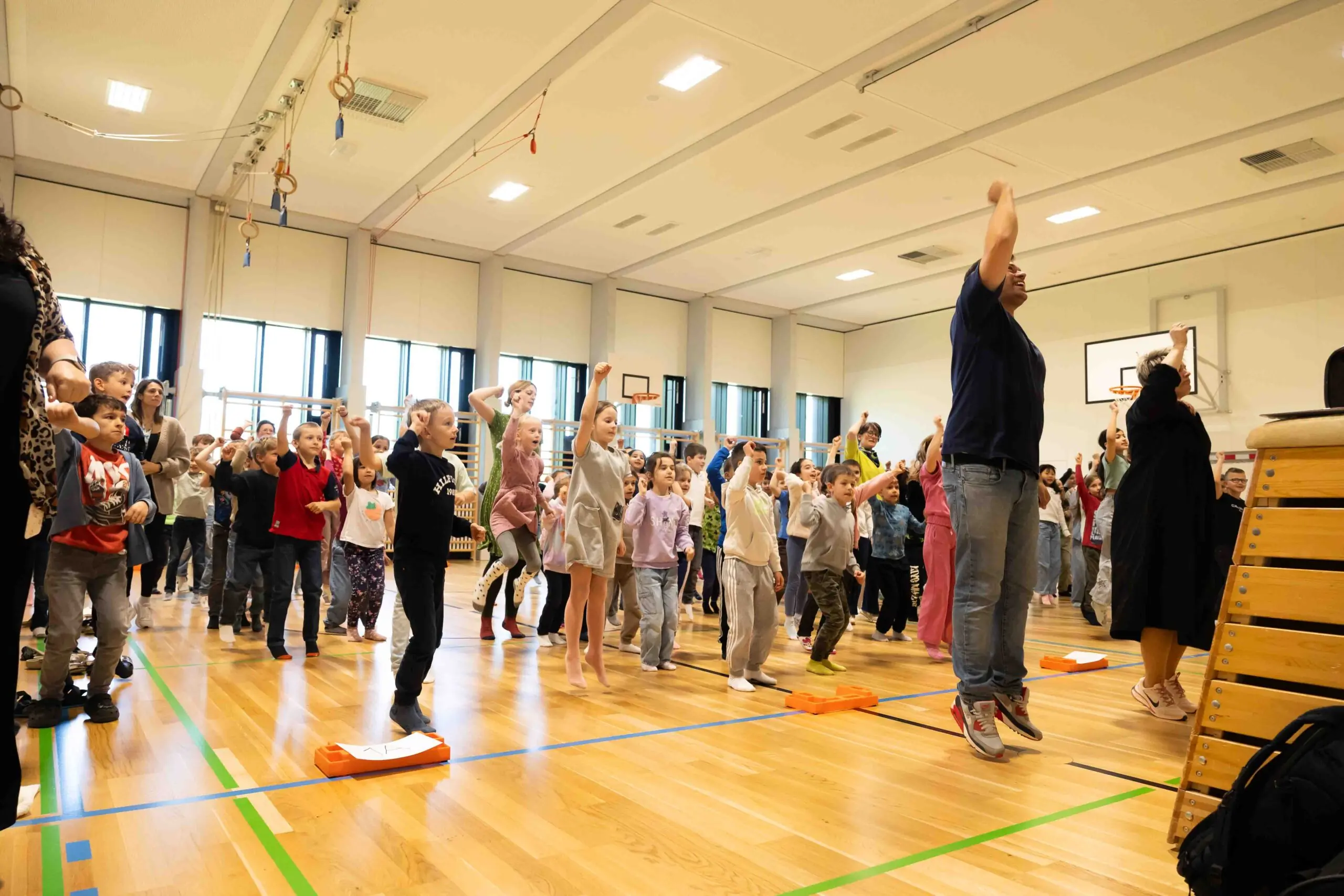 Eine Gruppe von Schulkindern springt gemeinsam mit ihrem Lehrer in einer Turnhalle nach oben – Training der Raumorientierung und motorischen Umsetzung.