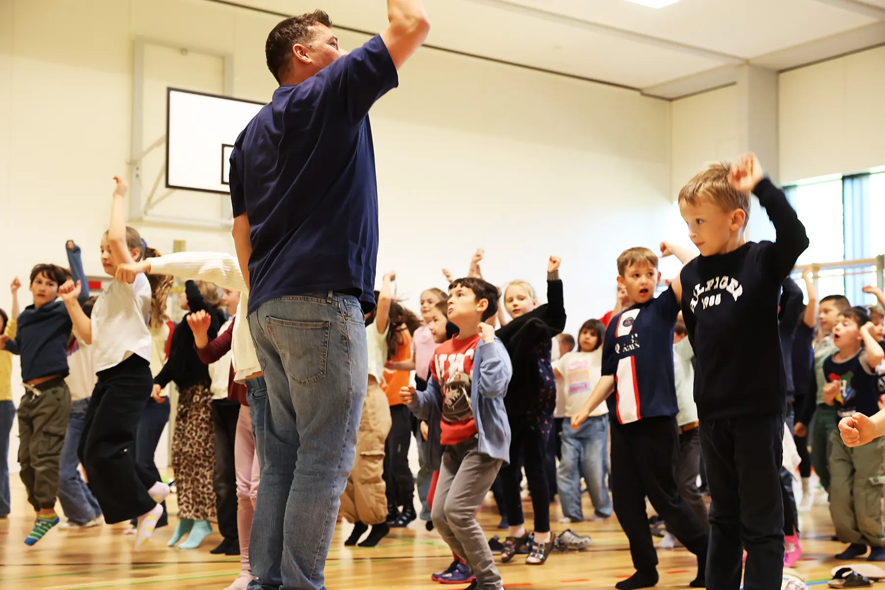 Eine Lehrkraft steht frontal vor einer Gruppe von Schulkindern in der Turnhalle und leitet einen Spiegeltanz an – direkte Nachahmung von Bewegungen.