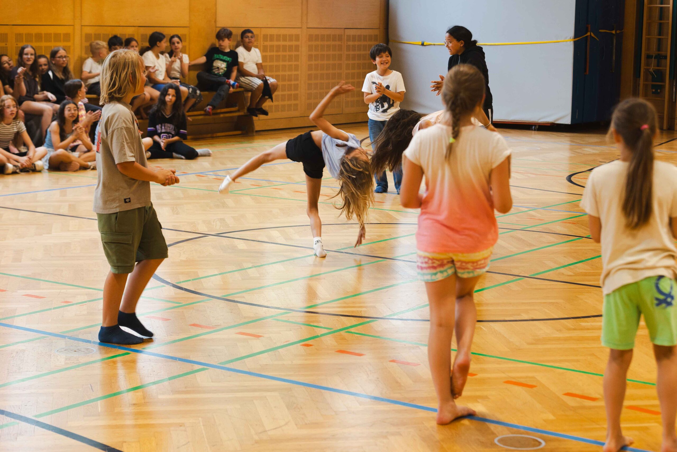 Eine Schülerin macht einen Radschlag in einer Turnhalle, während andere Kinder im Hintergrund zusehen – Symbol für individuelle Ausdruckskraft und Gemeinschaft.