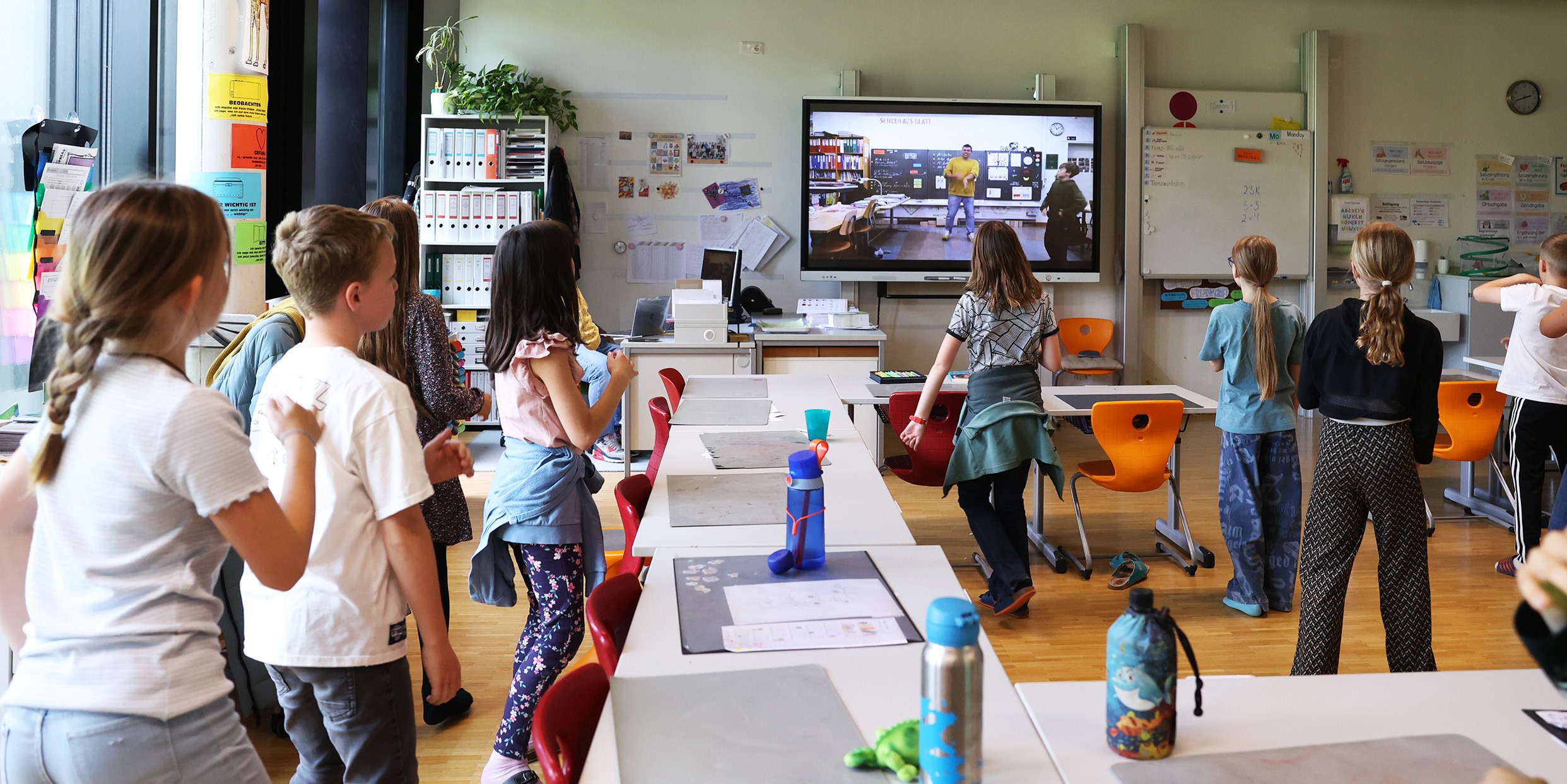Eine Schulklasse tanzt und bewegt sich im Klassenzimmer vor einem großen digitalen Bildschirm, auf dem ein groove2grow Video als staatlich geprüftes Unterrichtsmittel läuft.