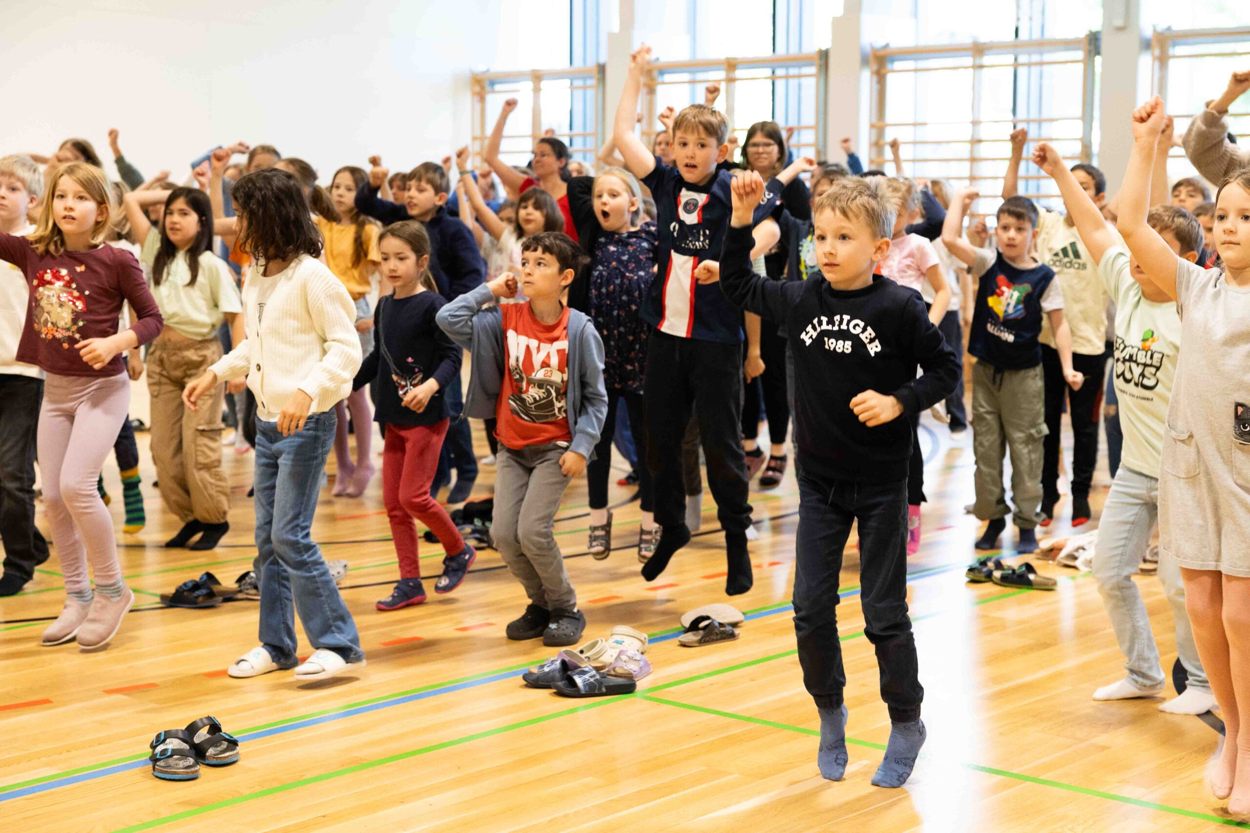 Eine ganze Volksschule springt und tanzt gemeinsam voller Energie in einer Turnhalle, um Synchronie und Kooperation durch das groove2grow Programm zu erleben.