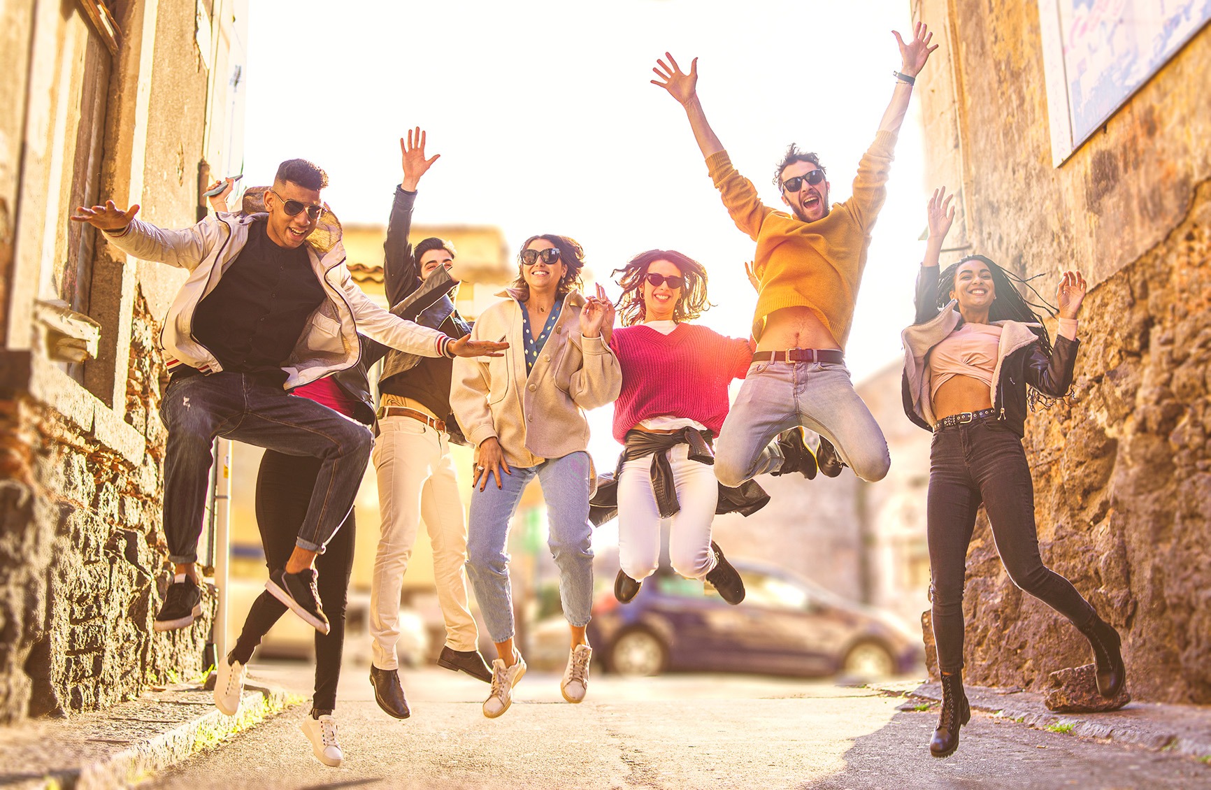 Eine vielfältige Gruppe junger Menschen springt und tanzt gemeinsam vor Freude in einer sonnigen Gasse – Symbol für sozialen Zusammenhalt.