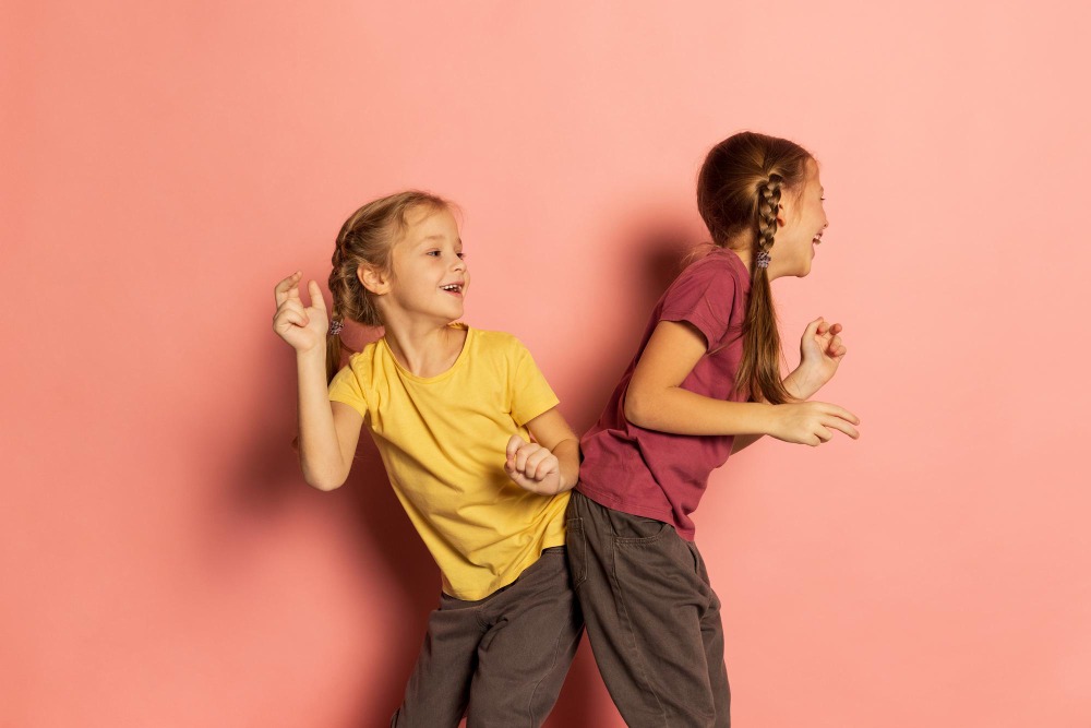Gefühle tänzerisch ausdrücken Zwei Mädchen tanzen lachend Rücken an Rücken vor rosa Hintergrund – Ausdruck von Freude und emotionaler Verbundenheit.