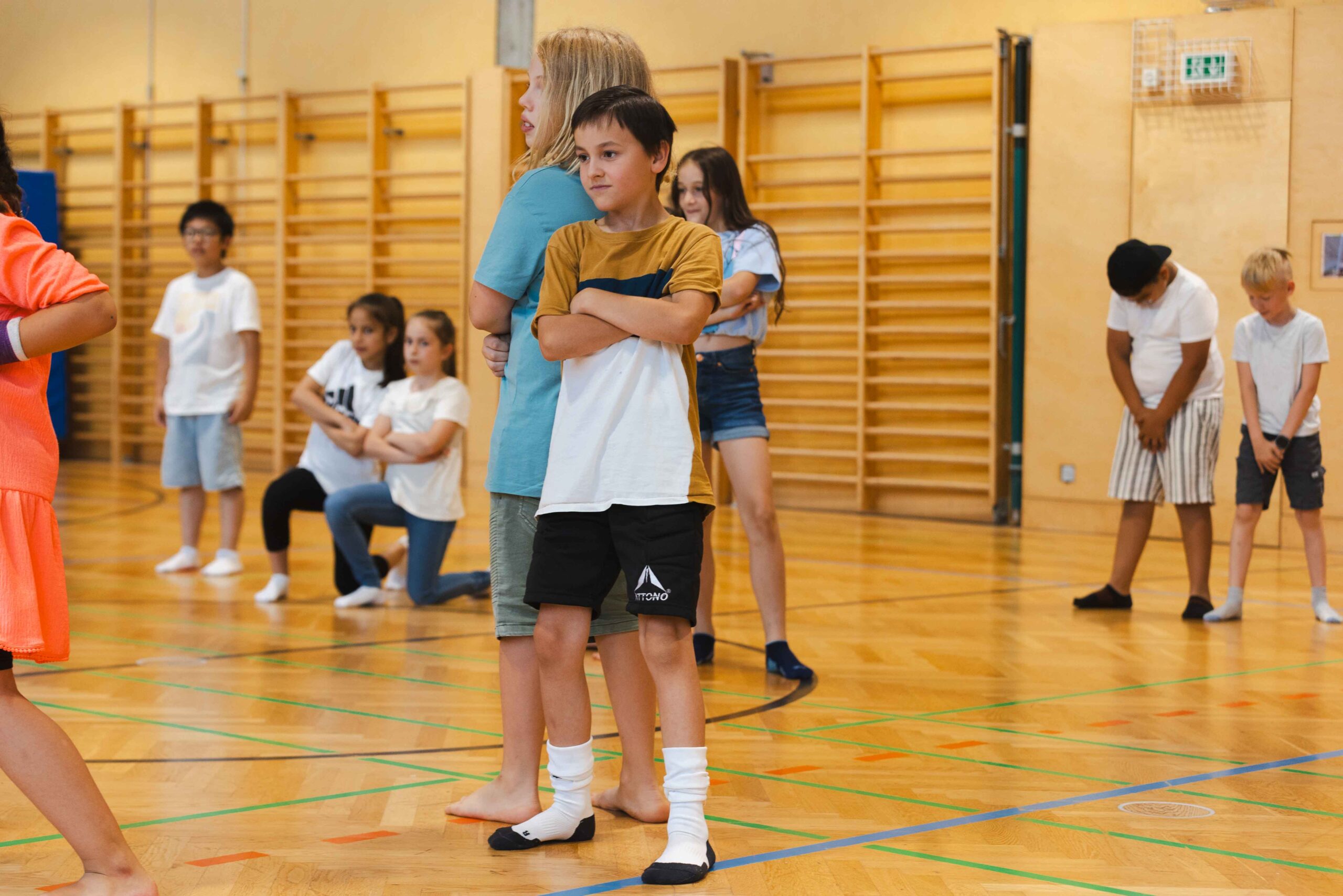Zwei Schüler stehen Rücken an Rücken mit verschränkten Armen in einer Turnhalle, während im Hintergrund andere Kinder zusehen – Ausdruck von Selbstbewusstsein und Teamgeist.