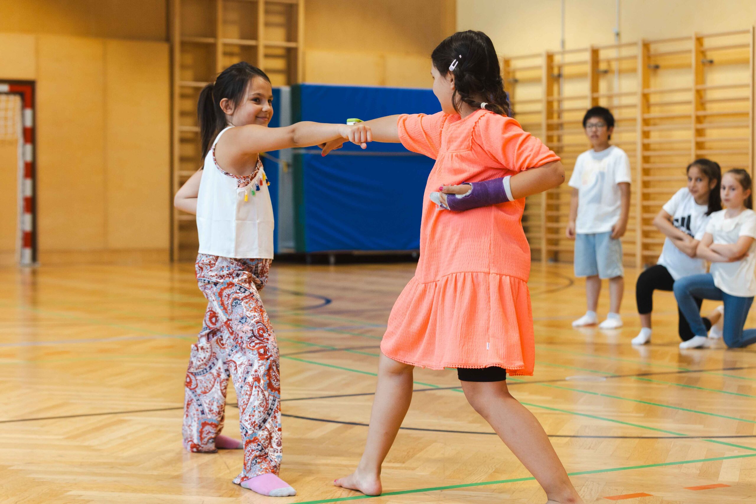 Zwei Schülerinnen in einer Turnhalle bei einer koordinierten Tanzbewegung – inspiriert vom Gemeinschaftsgeist kultureller Tanzbewegungen.